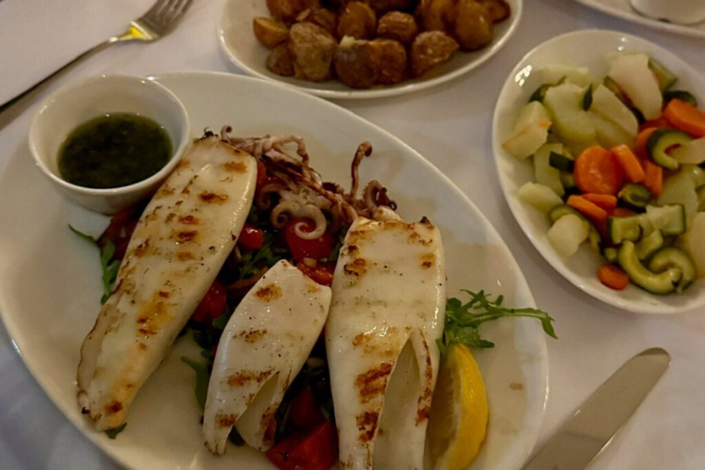 Gegrillter Tintenfisch mit Salat und Zitronenspalte, dazu Bratkartoffeln und Gemüse auf einem Restauranttisch.